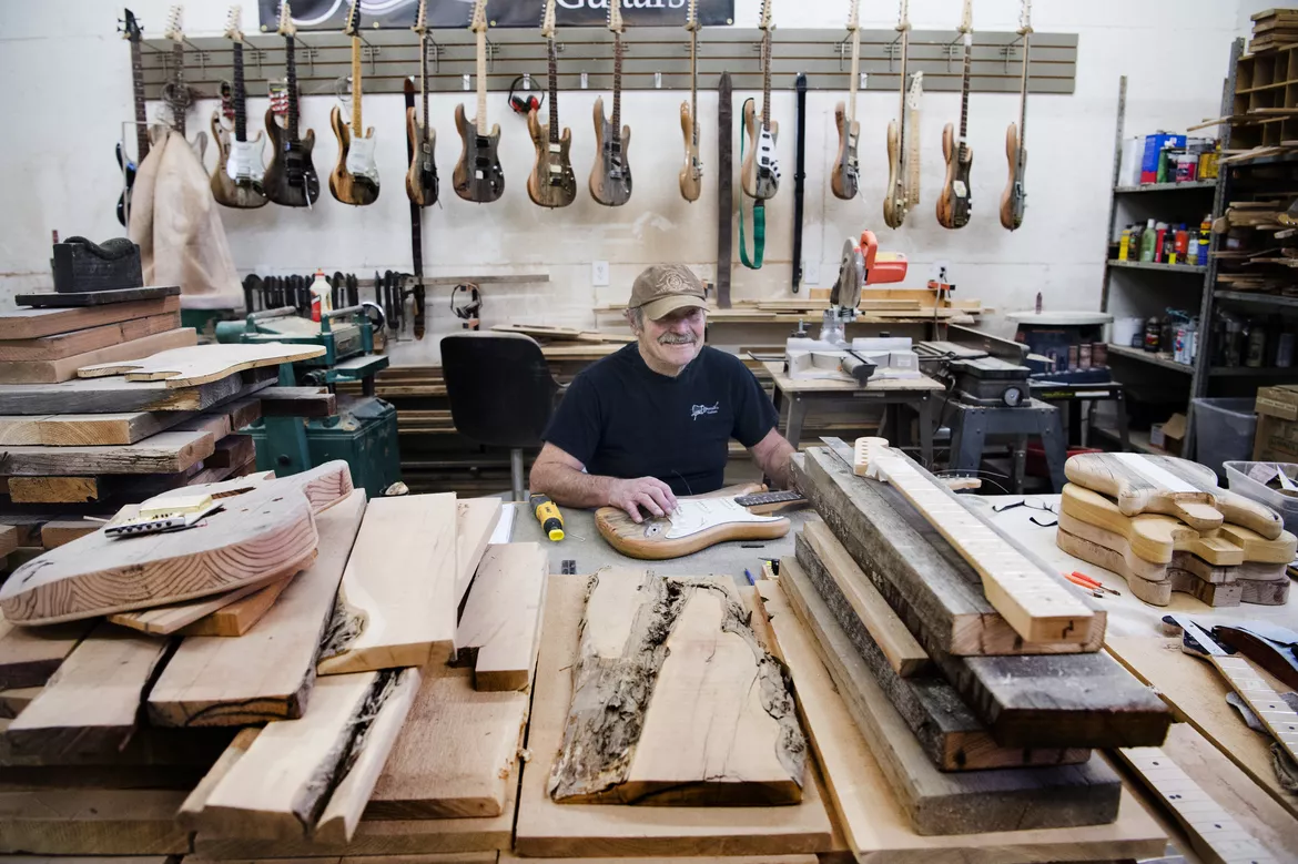 A photo of Lynn Ellsworth in his workshop, smiling at the camera and resting his hand over one of many guitars that are visble in the shot.