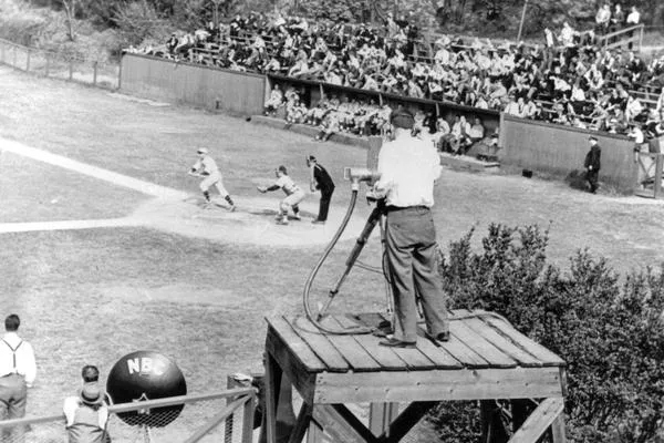 A photo taken of the first recording of a college baseball game! Several players play ball, under the foreground of a large camera recording them.