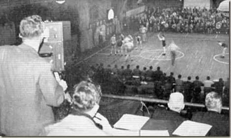 A poor quality photo showing a camera up in the press box of Fordham university, recording an active basketball game.
