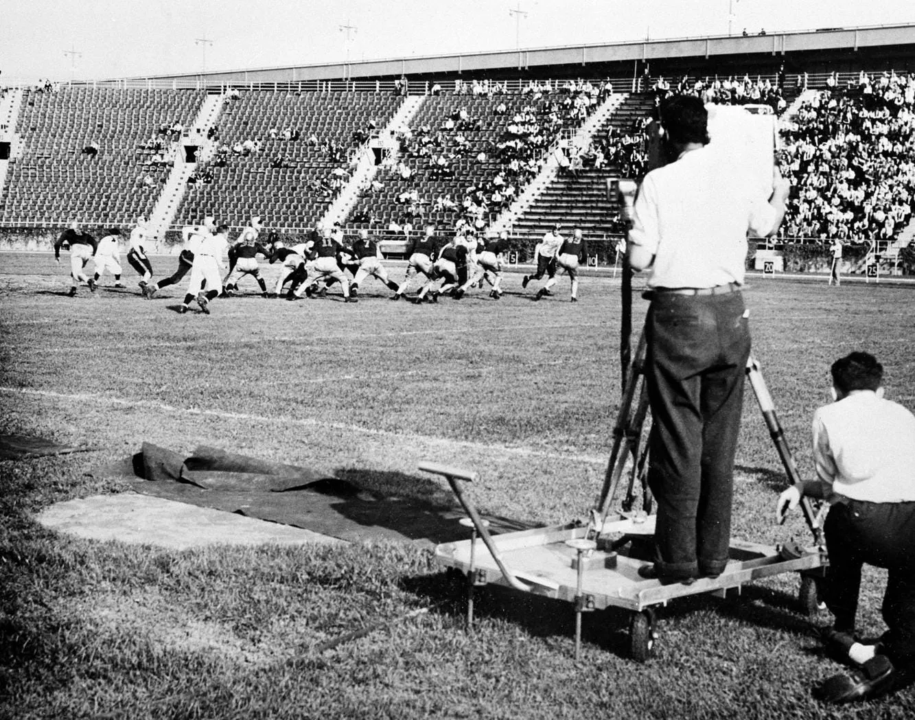 A photo from the period depicting a camera crow recording a set of footballers right as the game starts, with a modest crowd in the stands.