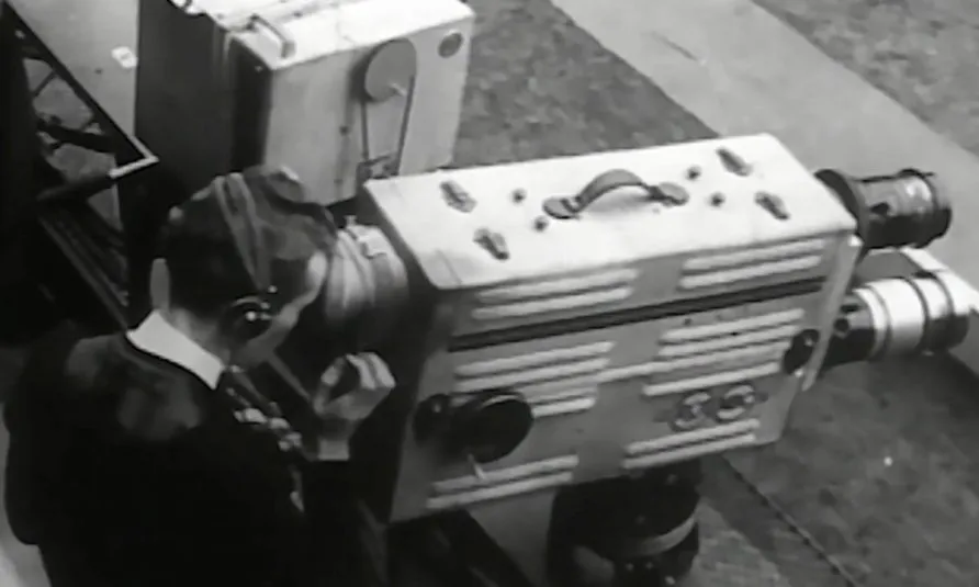 A close up of a camera operator operating   a complex tri-lensed camera of the 30s and recording a sports field.